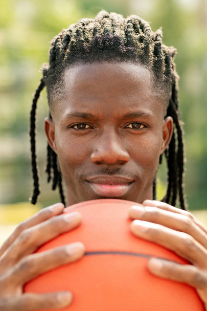 Joven jugador de baloncesto sosteniendo la pelota por la cara mientras está de pie cerca de la cámara y mirándote
 - Foto, Imagen