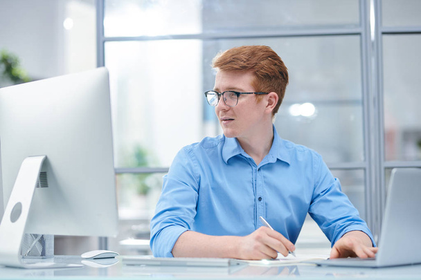 Молодой многозадачный офисный менеджер смотрит на экран компьютера, сидя перед ноутбуком и делая заметки
 - Фото, изображение