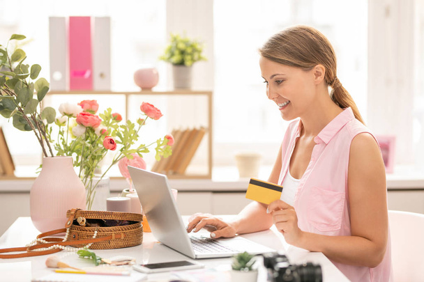 Młoda Wesoła kobieta z plastikową kartą patrząc na wyświetlacz laptopa podczas wprowadzania danych osobowych do zapłaty za zamówienie w sklepie internetowym - Zdjęcie, obraz