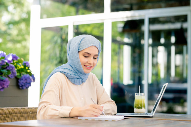 Joven estudiante musulmán en hijab haciendo notas de trabajo o haciendo tareas en casa al aire libre
 - Foto, Imagen