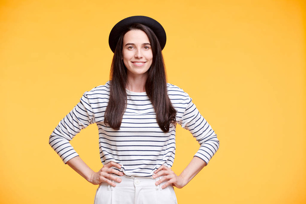 Young smiling woman in casualwear keeping her hands on waist while posing on yellow background in isolation - Foto, Imagem