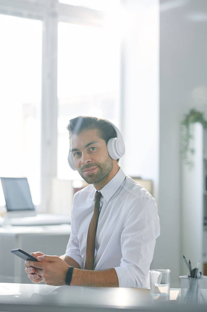 Trabalhador de escritório jovem confiante em fones de ouvido usando smartphone enquanto ouve música da lista de reprodução
 - Foto, Imagem