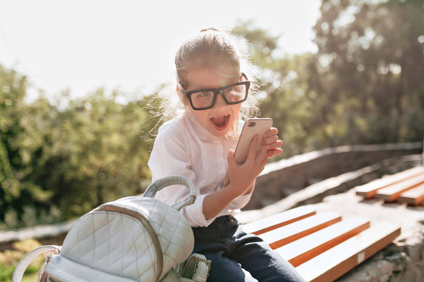 Zavři portrét legrační holčičky s brýlemi a bílou košilí, připravující se na studium a používání smartphone - Fotografie, Obrázek