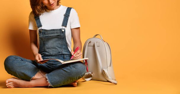 Portrait of a pensive girl in a white T-shirt and denim overalls, sitting on the floor with a red notebook and a pencil on a yellow background - Φωτογραφία, εικόνα