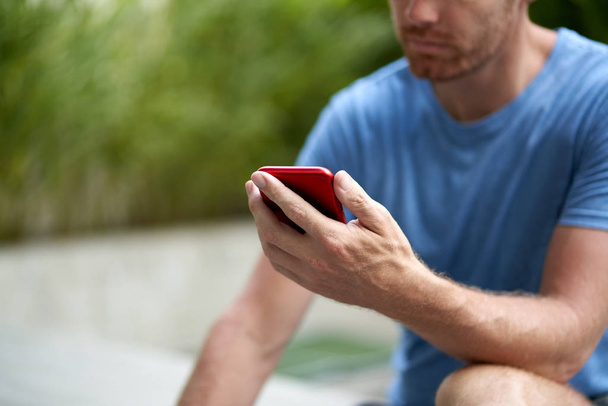 Candid lifestyle closeup shot of handsome with beard man sitting holding cellphone reading emails in blue athletic tshirt outdoors - Photo, Image