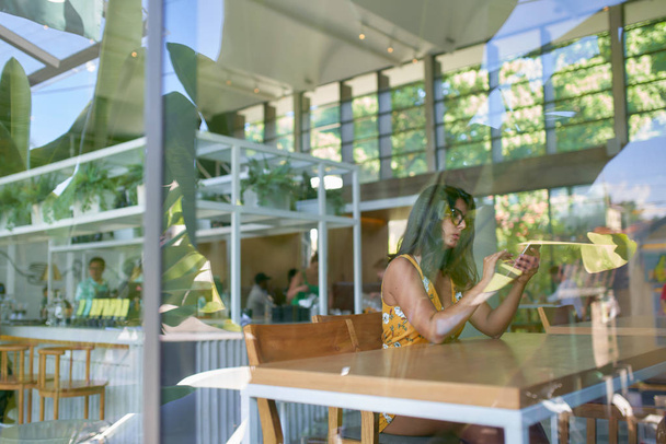 Lifestyle shot of fashionable hispanic millennial influencer typing on cellphone in modern trendy and bright coffee shop with plants - Photo, Image