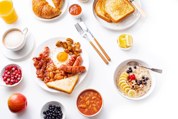 fried eggs with bacon and sausages with oatmeal and berries with fried toasts and jam on white background  - Foto, imagen
