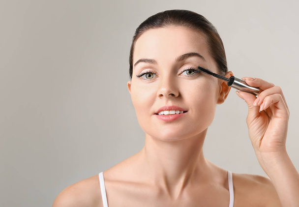 Beautiful young woman with eyelash extensions applying mascara against grey background - Zdjęcie, obraz