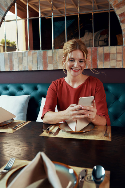 Belle jeune femme souriante en robe rouge vif assise à la table du restaurant et textos ou en utilisant une application mobile
 - Photo, image