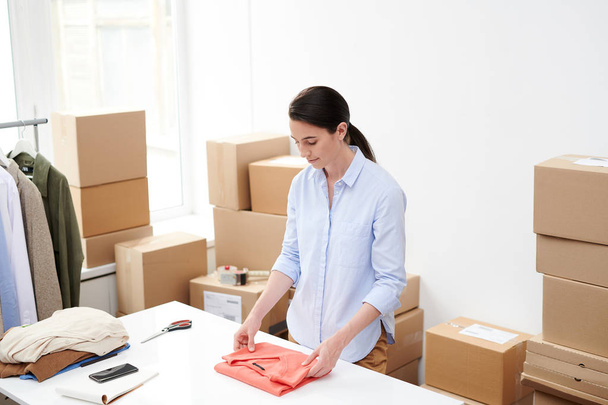 Young female online shop worker folding red pullover for packing while standing desk in office - Photo, Image