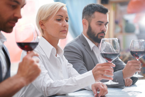 Serious blonde woman looking at red wine in bokal while evaluating color of the drink between her colleagues - Photo, Image