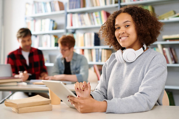 Menina multicultural sorridente casual em camisola olhando para você enquanto usa touchpad na sala de aula
 - Foto, Imagem