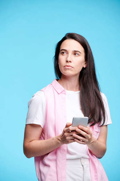 junge ernsthafte brünette Frau in Freizeitkleidung schaut beiseite, während sie in der Isolation im Smartphone blättert - Foto, Bild