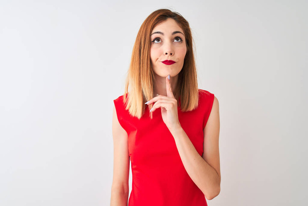 Redhead zakenvrouw het dragen van elegante rode jurk staande over geïsoleerde witte achtergrond denken geconcentreerd over twijfel met de vinger op de kin en opzoeken benieuwd - Foto, afbeelding