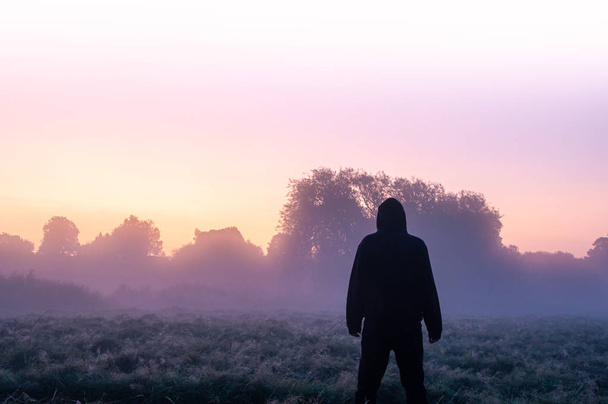 神秘的な孤独なフード付き、美しい早朝の霧の朝の野原に立って、日の出を見て. - 写真・画像