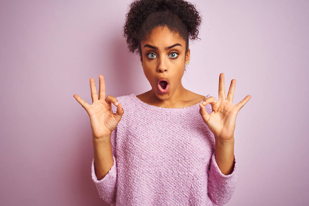 Joven mujer afroamericana vistiendo suéter de invierno de pie sobre el fondo rosa aislado mirando sorprendido y sorprendido haciendo símbolo de aprobación ok con los dedos. Expresión loca
 - Foto, Imagen