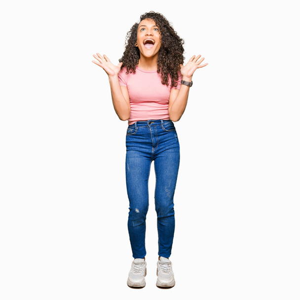Young beautiful woman with curly hair wearing pink t-shirt celebrating crazy and amazed for success with arms raised and open eyes screaming excited. Winner concept - Fotografie, Obrázek