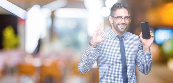 Young business man showing smartphone screen over isolated background doing ok sign with fingers, excellent symbol - Foto, Bild