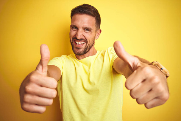 Молодой красивый мужчина в повседневной желтой футболке на желтом изолированном фоне, одобряя позитивный жест рукой, улыбается и радуется успеху. Жест победителя
. - Фото, изображение