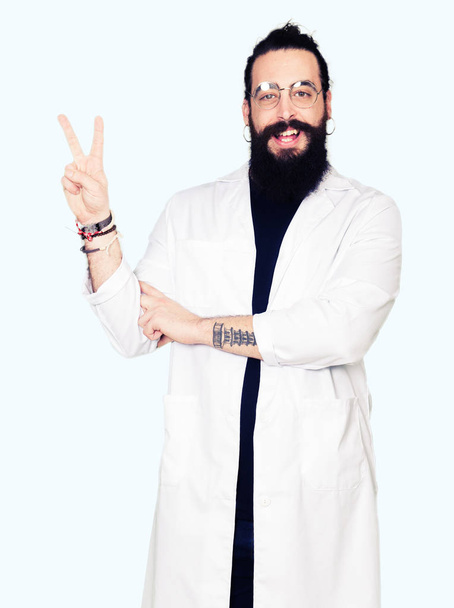 Doctor therapist man with long hair and bear wearing white coat smiling with happy face winking at the camera doing victory sign. Number two. - 写真・画像