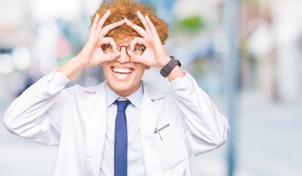Joven científico guapo con gafas haciendo un buen gesto como binoculares sacando la lengua, ojos mirando a través de los dedos. Expresión loca
. - Foto, imagen