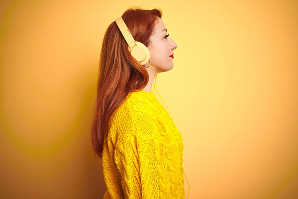 横を見て黄色の孤立した背景の上にヘッドフォンを使用して音楽を聴く若い赤毛の女性は、自信のある笑顔で自然な顔でプロフィールポーズをリラックス. - 写真・画像