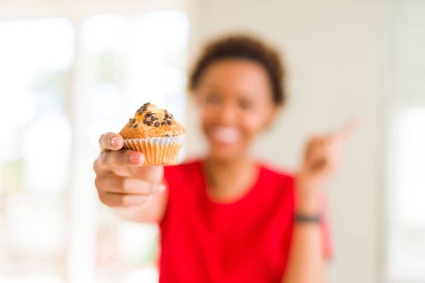 Young african american woman eating chocolate chips muffins very happy pointing with hand and finger to the side - Фото, зображення