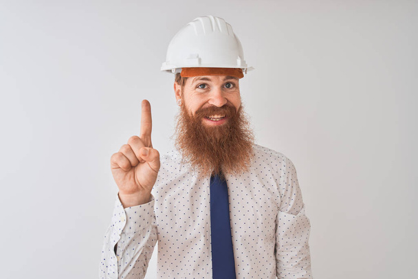 Νεαρή κοκκινομάλλα Ιρλανδή αρχιτέκτονας άντρας φορώντας κράνος ασφαλείας πάνω από απομονωμένο λευκό φόντο δείχνοντας και δείχνοντας με το δάχτυλο νούμερο ένα, χαμογελώντας σίγουροι και χαρούμενοι. - Φωτογραφία, εικόνα