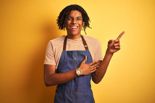 Homem barista afro-americano com dreadlocks vestindo avental sobre fundo amarelo isolado com um grande sorriso no rosto, apontando com a mão e o dedo para o lado olhando para a câmera
. - Foto, Imagem