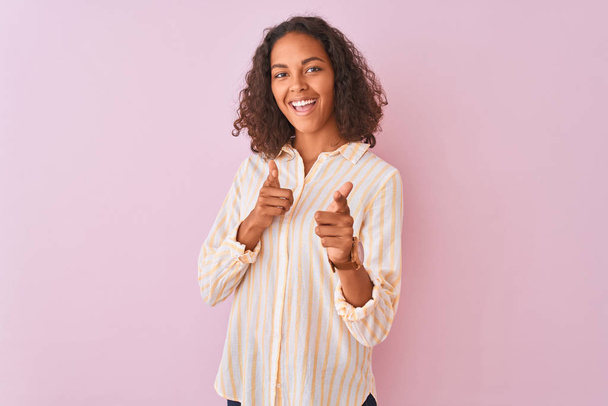 孤立したピンクの背景の上に立つストライプのシャツを着た若いブラジル人女性は、幸せで面白い顔でカメラに指を指しています。良いエネルギーとバイブ. - 写真・画像