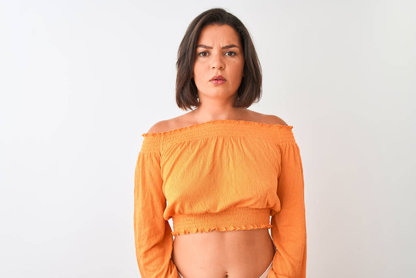Młoda piękna kobieta ubrana w pomarańczowy t-shirt stojący nad białym tłem z poważnym wyrazem na twarzy. Proste i naturalne patrząc na kamerę. - Zdjęcie, obraz