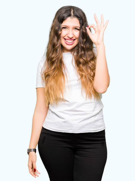 若くてきれいな女性の手と指で ok サインをしている肯定的な笑みを浮かべてカジュアルな白い t シャツを身に着けています。成功した式. - 写真・画像