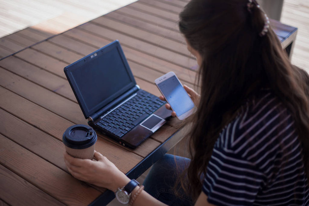Brunette girl with a large wrist watch, half-turned, works at a laptop standing on table. Nearby is a smartphone. - Foto, afbeelding