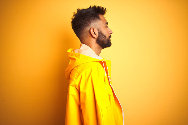 Young indian man wearing raincoat standing over isolated yellow background looking to side, relax profile pose with natural face with confident smile. - Photo, Image