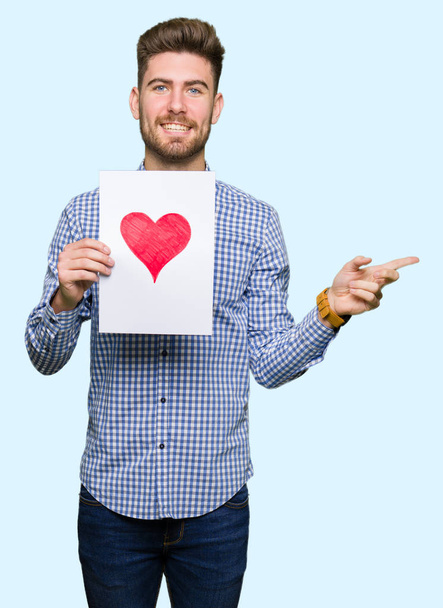 Young handsome man holding paper with red heart very happy pointing with hand and finger to the side - Foto, immagini