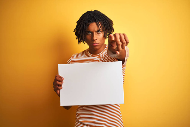 Afro amerykański człowiek z dredy trzyma banner na białym tle żółte wskazując palcem do aparatu i do Ciebie, znak strony, pozytywny i pewny gest od przodu - Zdjęcie, obraz