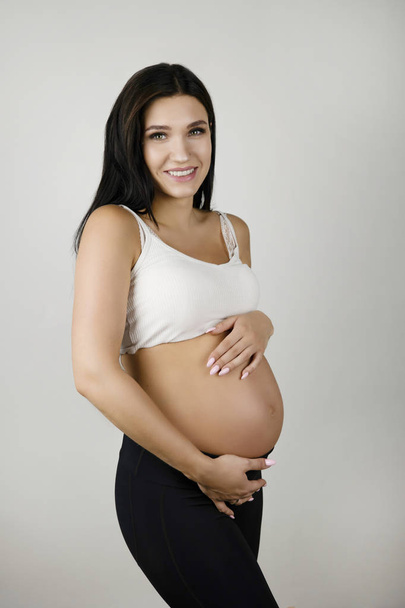 beautiful pregnant brunette woman holding her pregnant naked belly looking content on isolated white background - Foto, afbeelding