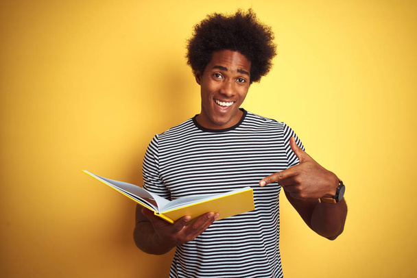 Afro american student man reading book standing over isolated yellow background very happy pointing with hand and finger - Фото, изображение