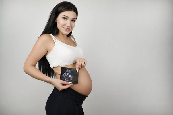 beautiful pregnant brunette woman holding ultrasound picture of her baby feeling love smiling on isolated white background - Photo, Image