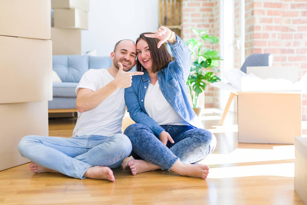 Young couple sitting on the floor arround cardboard boxes moving to a new house smiling making frame with hands and fingers with happy face. Creativity and photography concept. - Photo, Image
