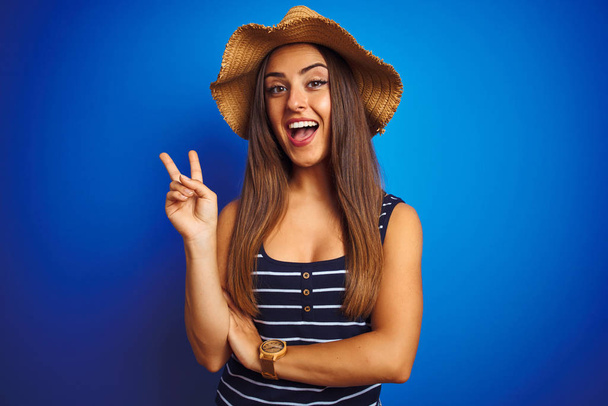 Jeune belle femme portant un t-shirt rayé et un chapeau d'été sur fond bleu isolé souriant avec un clin d'oeil heureux au visage de la caméra faisant signe de victoire. Numéro deux
. - Photo, image