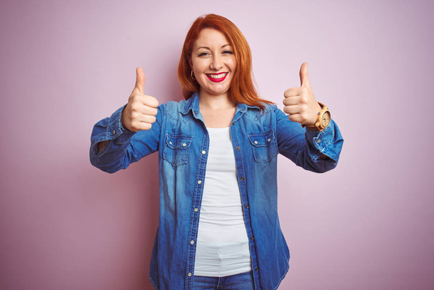 Youg beautiful redhead woman wearing denim shirt standing over isolated pink background success sign doing positive gesture with hand, thumbs up smiling and happy. Cheerful expression and winner gesture. - Photo, Image