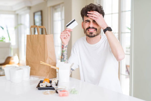 Młody mężczyzna jedzenie sushi azjatycki z dostawa do domu za pomocą karty kredytowej jako płatności podkreślił z ręki na głowie, w szoku ze wstydu i niespodzianka twarzy, gniew i frustrację. Strach i zdenerwowany za błąd. - Zdjęcie, obraz