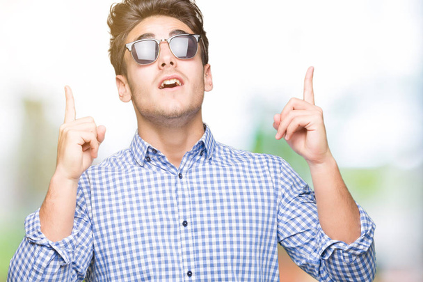 Jonge knappe man dragen van een zonnebril op geïsoleerde achtergrond verbaasd en verrast opzoeken en wijzen met vingers en opgeheven armen. - Foto, afbeelding