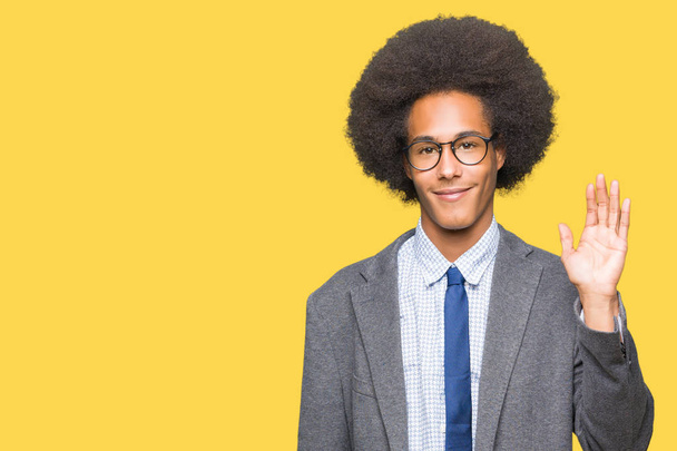 Jovem homem de negócios afro-americano com cabelo afro usando óculos Waiving dizendo olá feliz e sorridente, gesto de boas-vindas amigável
 - Foto, Imagem