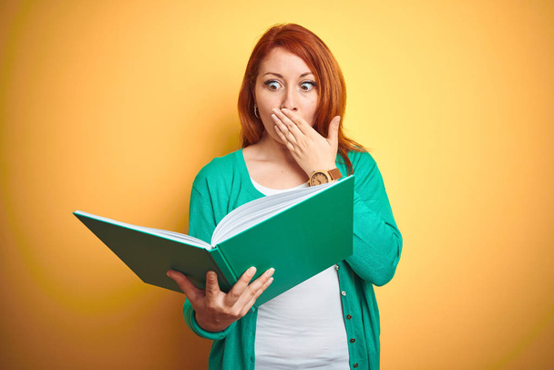 Young redhead student woman reading green book over yellow isolated background cover mouth with hand shocked with shame for mistake, expression of fear, scared in silence, secret concept - Foto, Bild