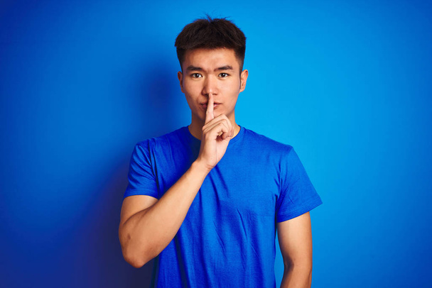 Jonge Aziatische Chinese man draagt t-shirt staande over geïsoleerde blauwe achtergrond vragen om stil te zijn met de vinger op de lippen. Stilte en geheim concept. - Foto, afbeelding