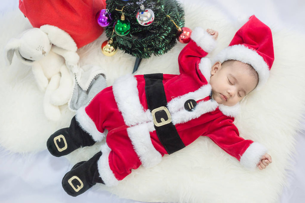 little baby wearing Santa Claus costume sleep on white fur carpet with Christmas tree. Concept of celebrates Christmas and New Year's holidays. - Photo, image