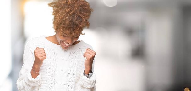 Beautiful young african american woman wearing winter sweater over isolated background very happy and excited doing winner gesture with arms raised, smiling and screaming for success. Celebration concept. - Zdjęcie, obraz