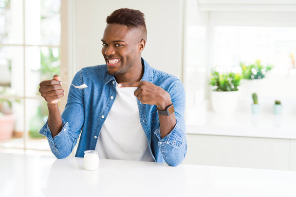 Afrikkalainen amerikkalainen mies syö terveellistä luonnollista jogurttia lusikalla erittäin onnellinen osoittaa kädellä ja sormella
 - Valokuva, kuva